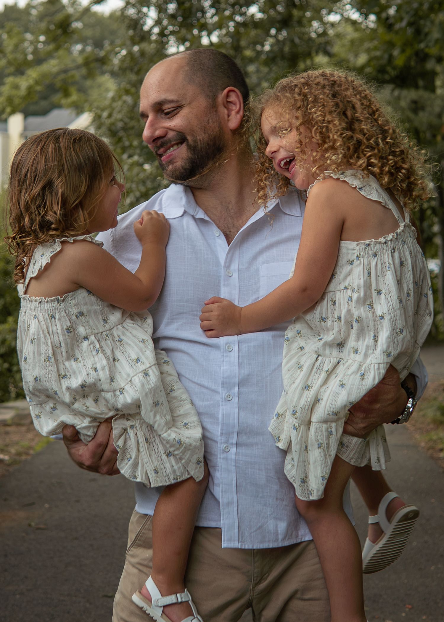 A smiling bald man holds two young girls in matching white dresses outdoors, sharing a joyful family moment.