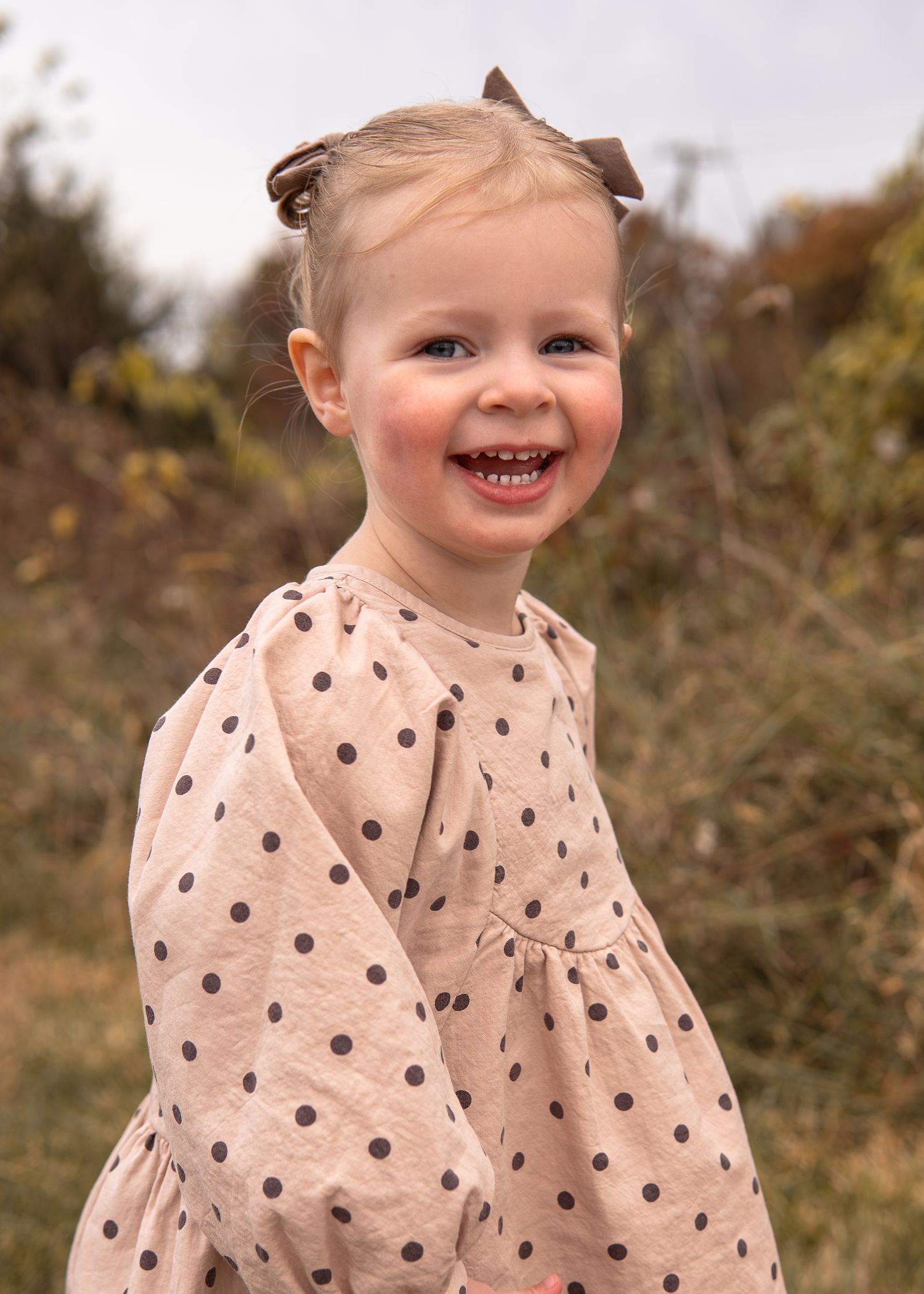 Cheerful toddler girl with a brown bow in her hair, wearing a pink polka dot dress, smiling outdoors.