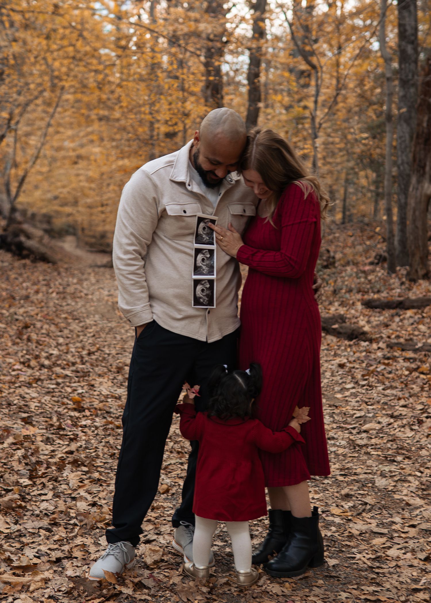 Family of three sharing emotional moment with ultrasound photos in autumn forest setting.