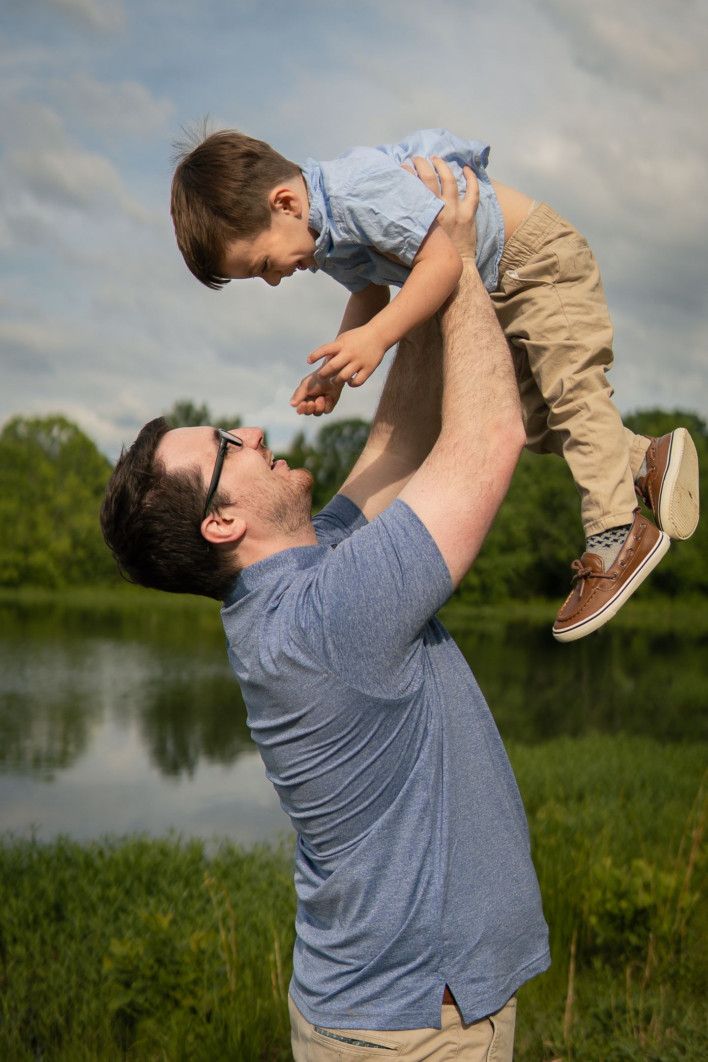 A playful moment captured by a lake as an adult lifts a child high into the air against a cloudy sky.