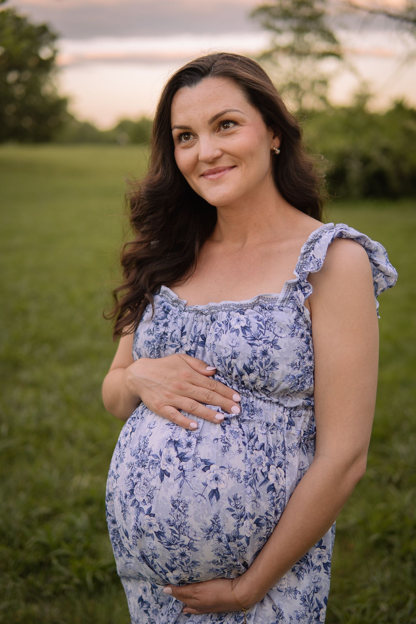 A woman in a blue and white floral dress poses outdoors in a grassy field during golden hour.