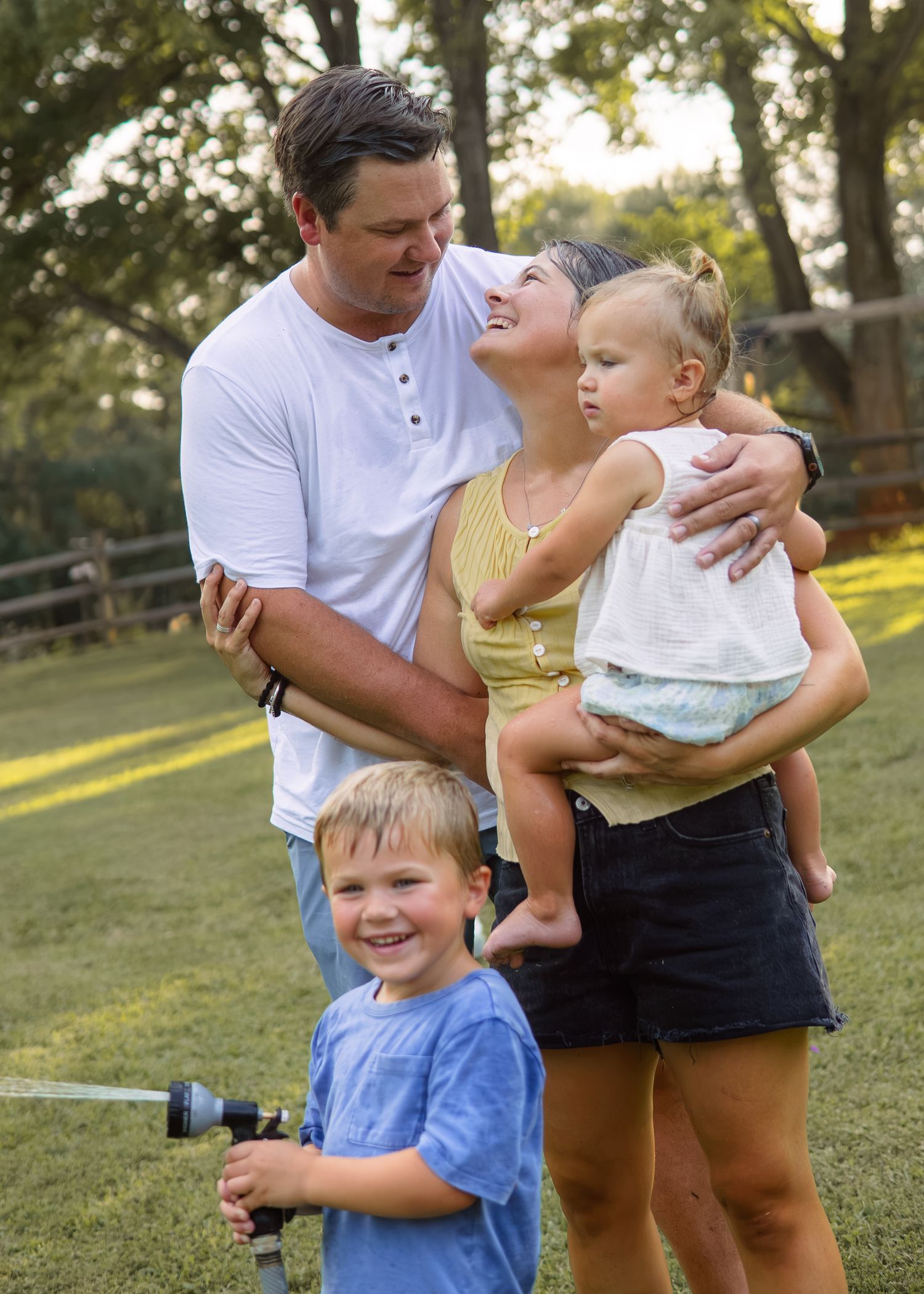 A smiling family with two young children poses together outdoors near a garden sprinkler.