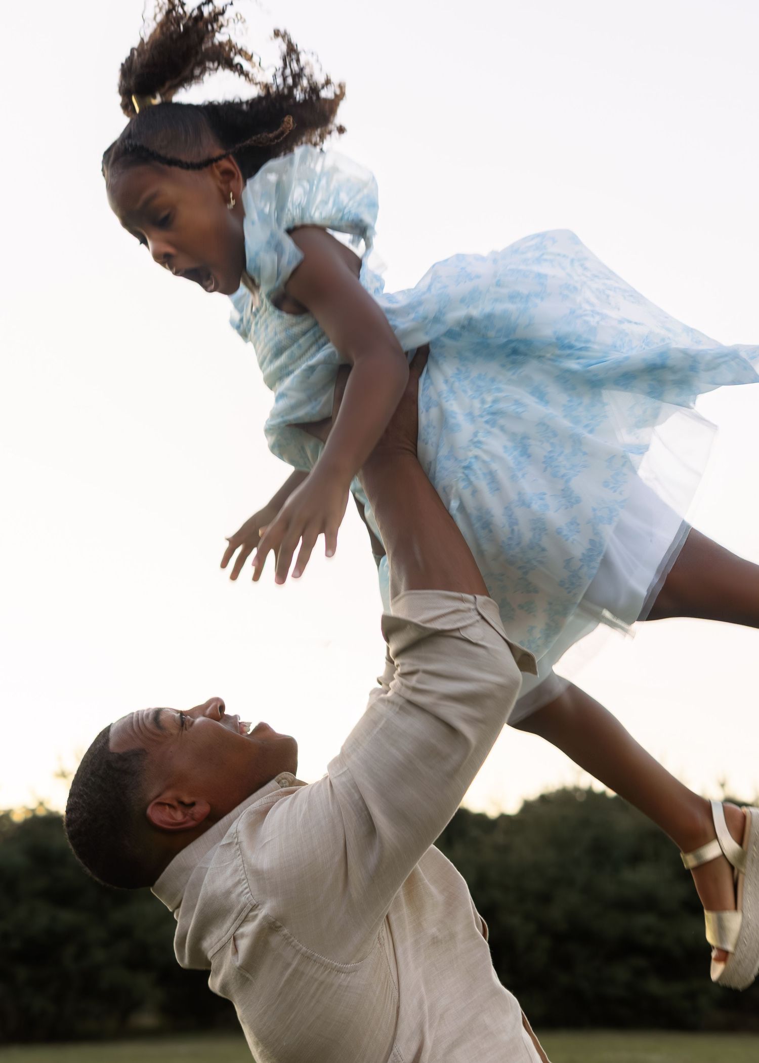 A father joyfully lifts his young daughter high in the air during a playful outdoor family portrait.