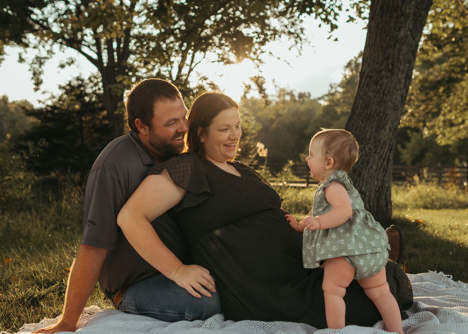 Pregnant woman sits with husband and baby outdoors in golden hour light near a tree on a blanket.
