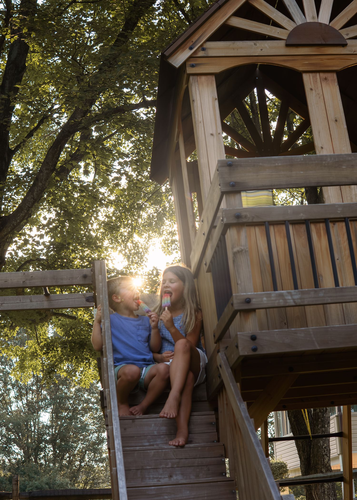 Two children sit on wooden treehouse slide enjoying popsicles in golden sunset light.