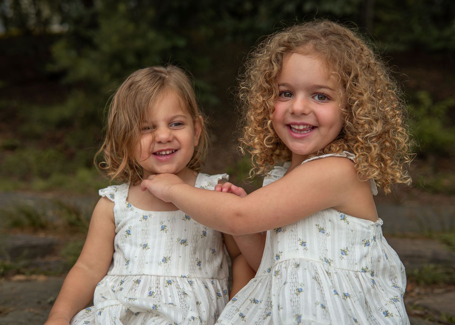 Two smiling young girls in matching white floral dresses pose together outdoors in a warm, natural setting.