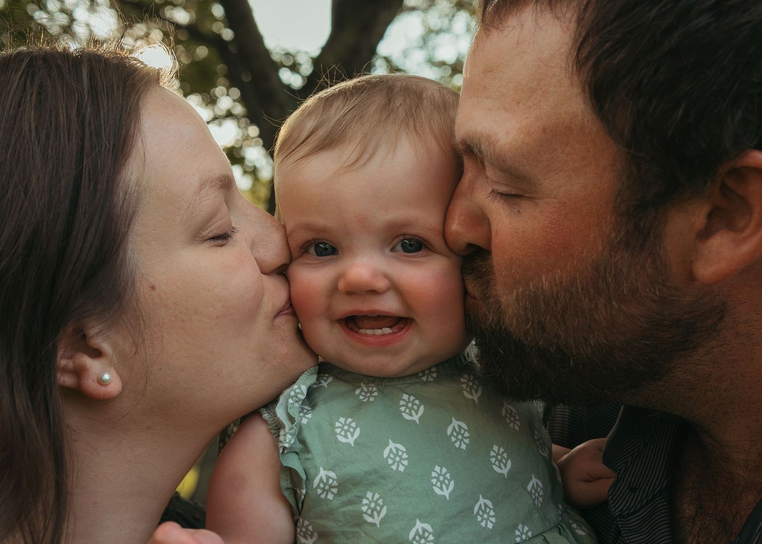 Two parents kissing their smiling baby on the cheeks outdoors in a heartwarming family portrait.