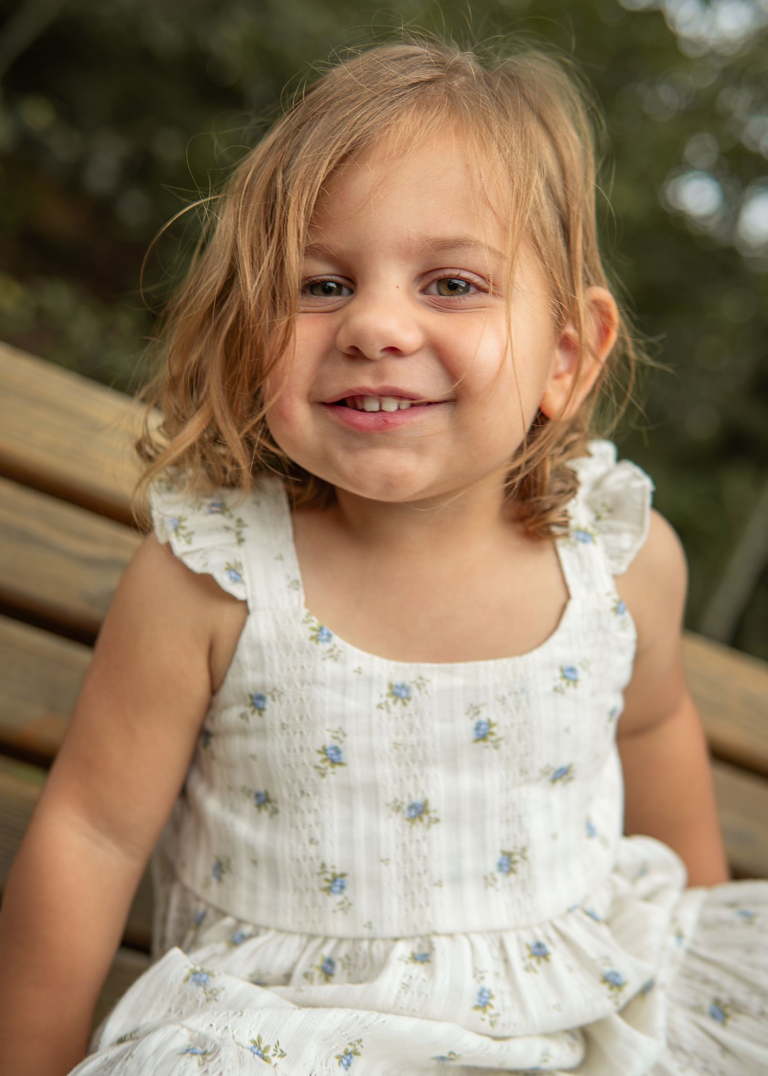 Smiling toddler girl with curly blonde hair wearing a white floral sundress sitting outdoors.