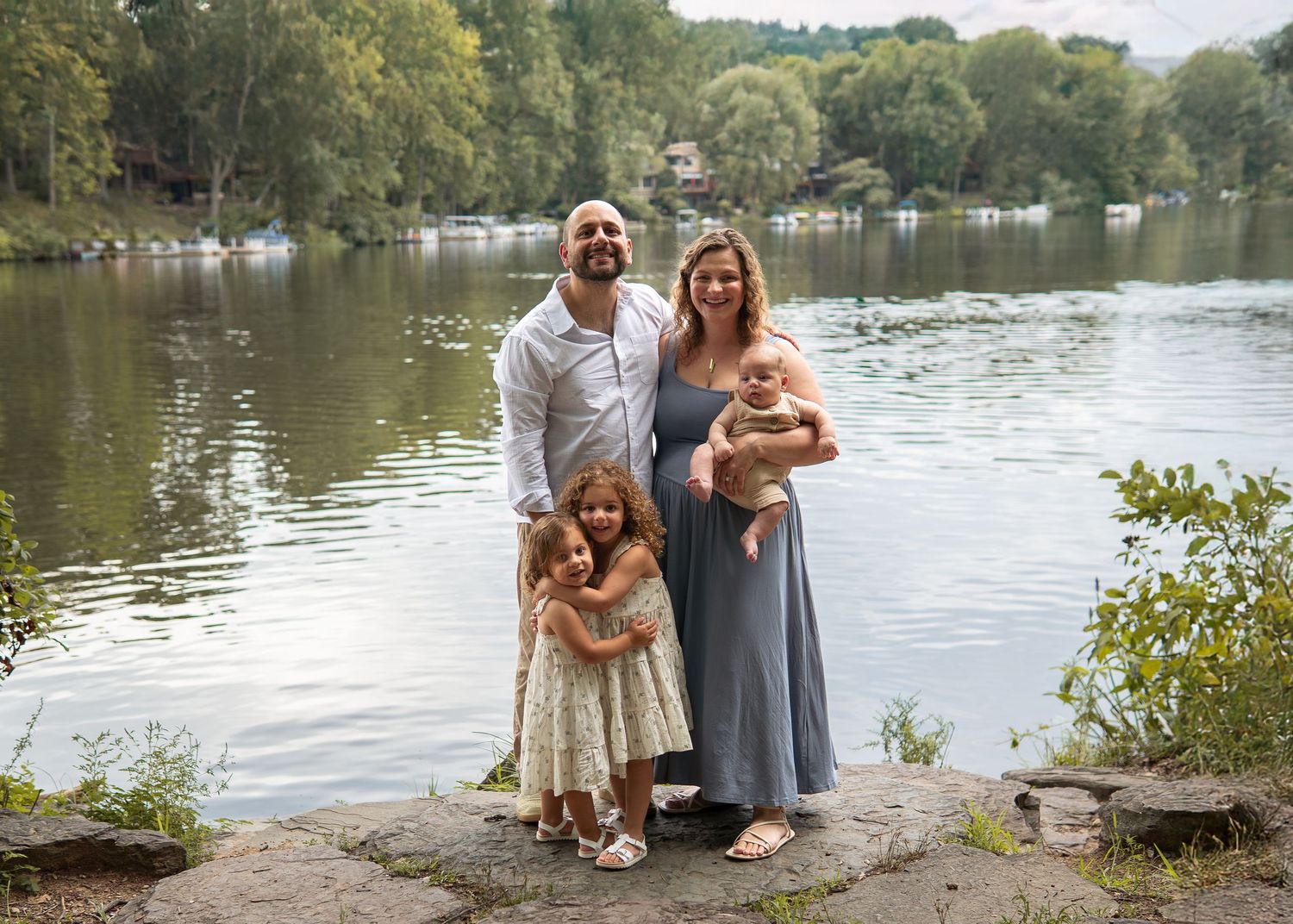 Family of four posing by a serene lake, parents holding children, surrounded by lush greenery on a cloudy day.