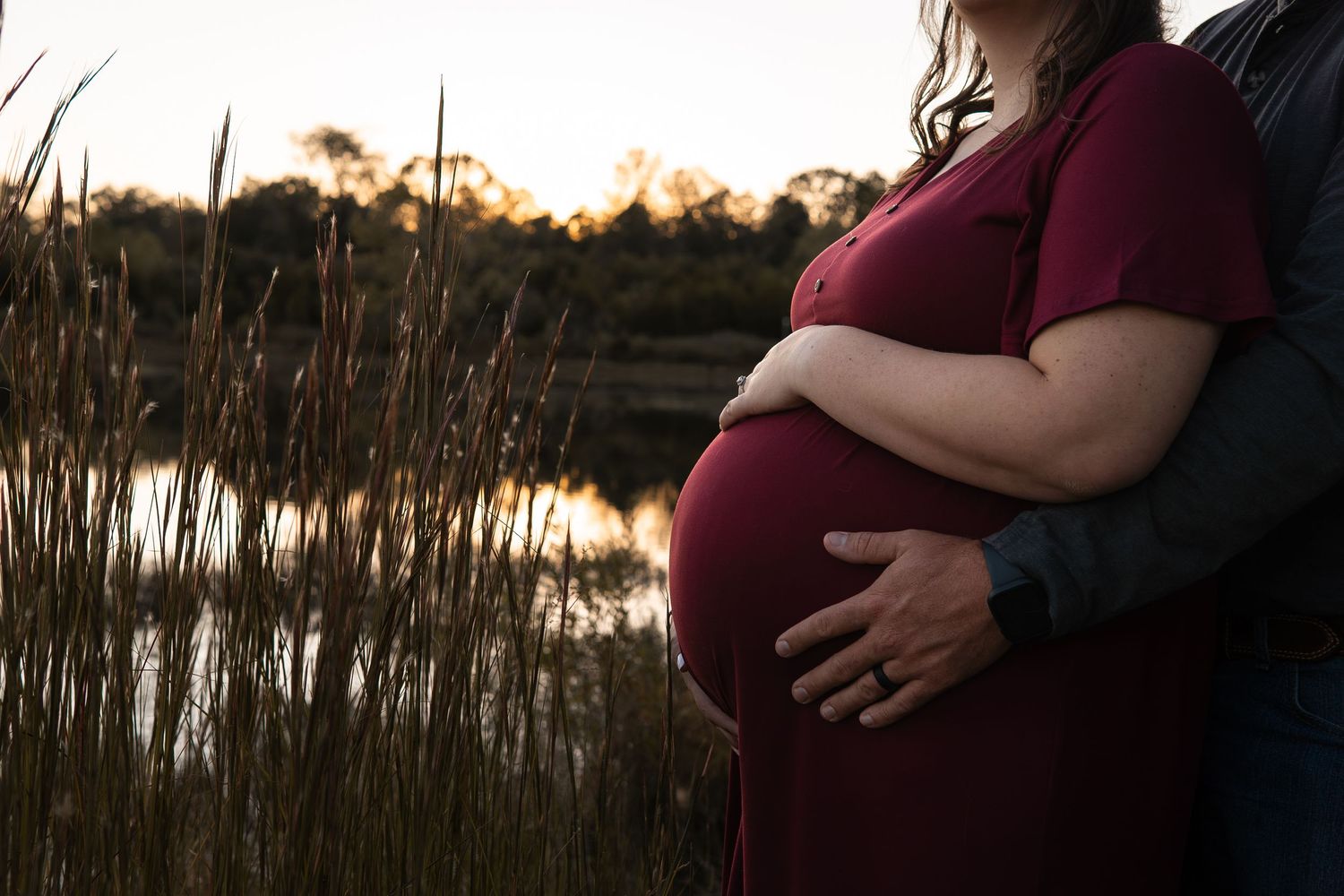 Pregnant woman in red dress cradling baby bump outdoors near a reflective pond at golden hour sunset.