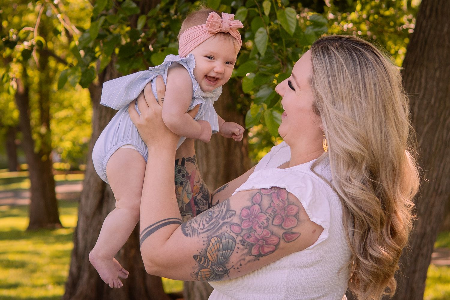 A playful moment between parent and baby wearing a pink bow in a sunlit park setting with trees.