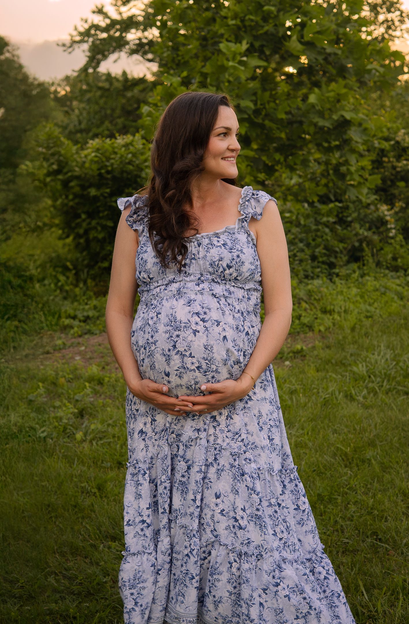 A floral maternity dress worn in a lush garden setting during golden hour with hands cradling the baby bump.