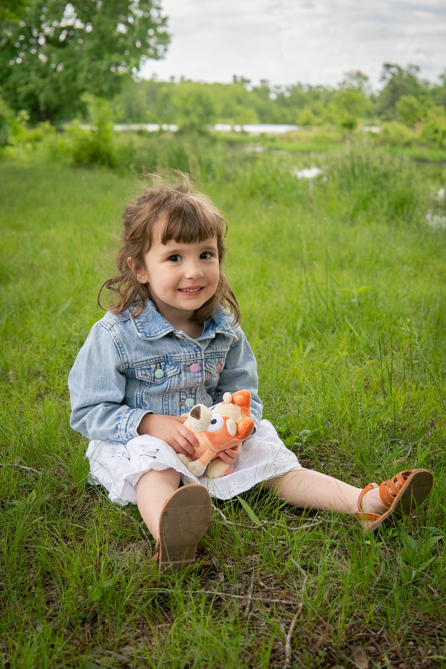 A small child in denim jacket and white dress sits in green grass while playing with a toy.
