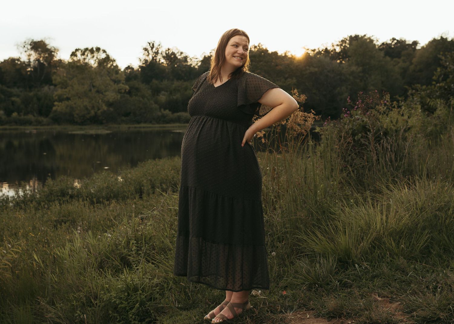 A pregnant woman in a black maxi dress standing near a pond at golden hour.