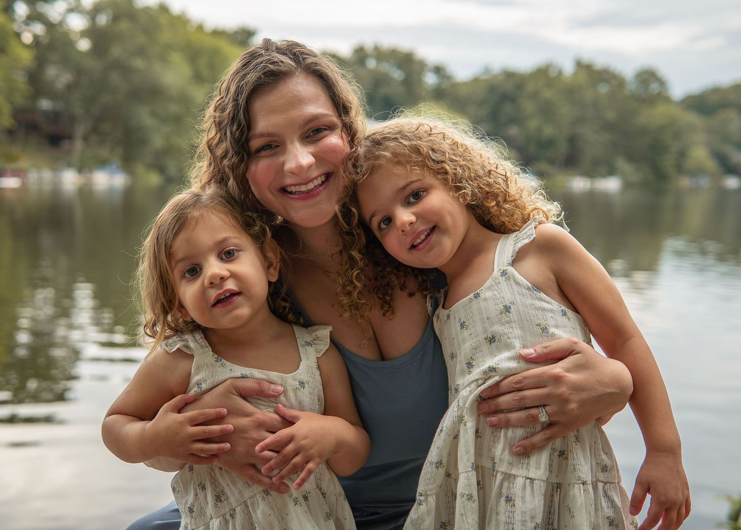 A smiling woman hugs two young girls in floral dresses near a serene lake surrounded by lush green trees.