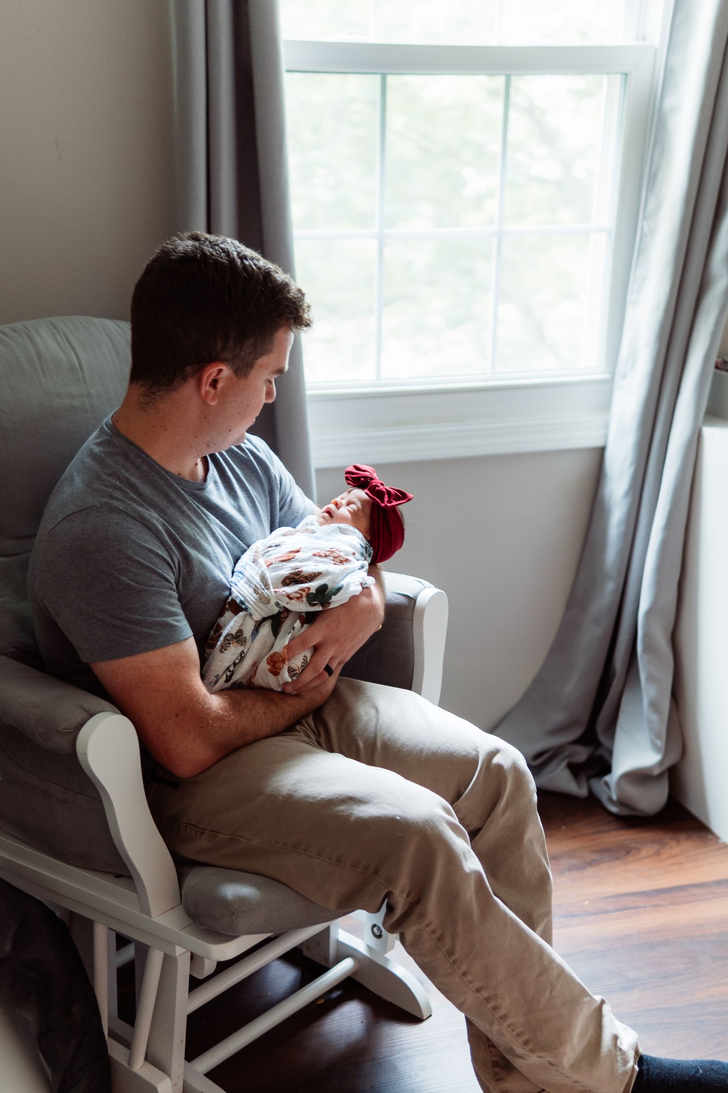 A person sits in a rocking chair holding a swaddled newborn near a window.