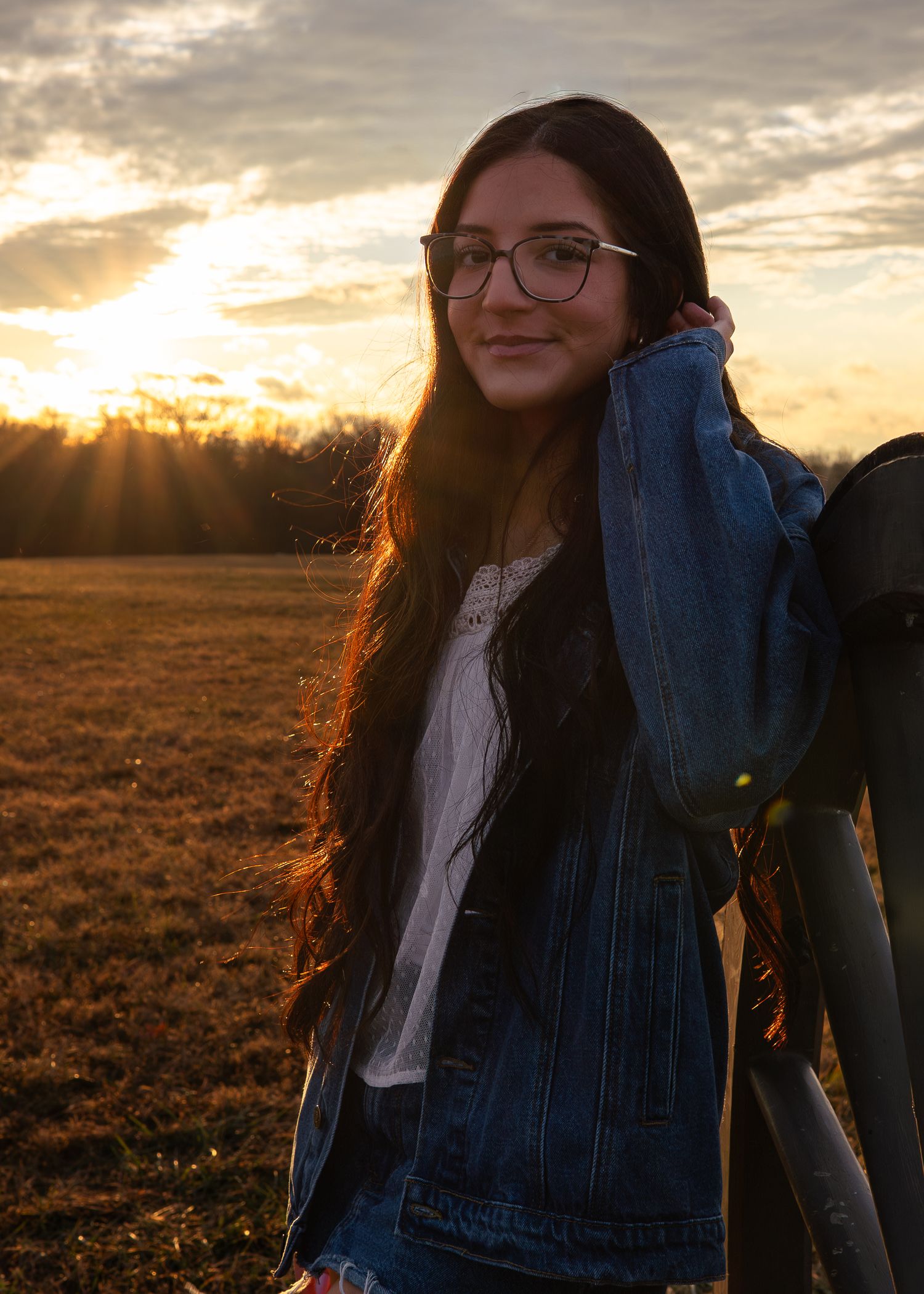 Smiling young woman with glasses in denim jacket posing in golden field at sunset.