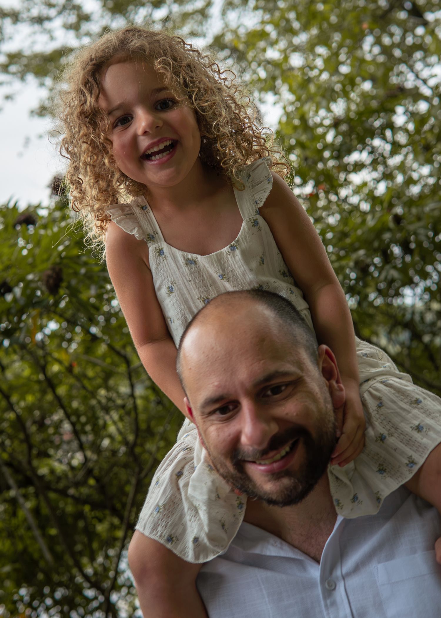Smiling bald father with curly-haired little girl sitting on his shoulders outdoors among green trees.