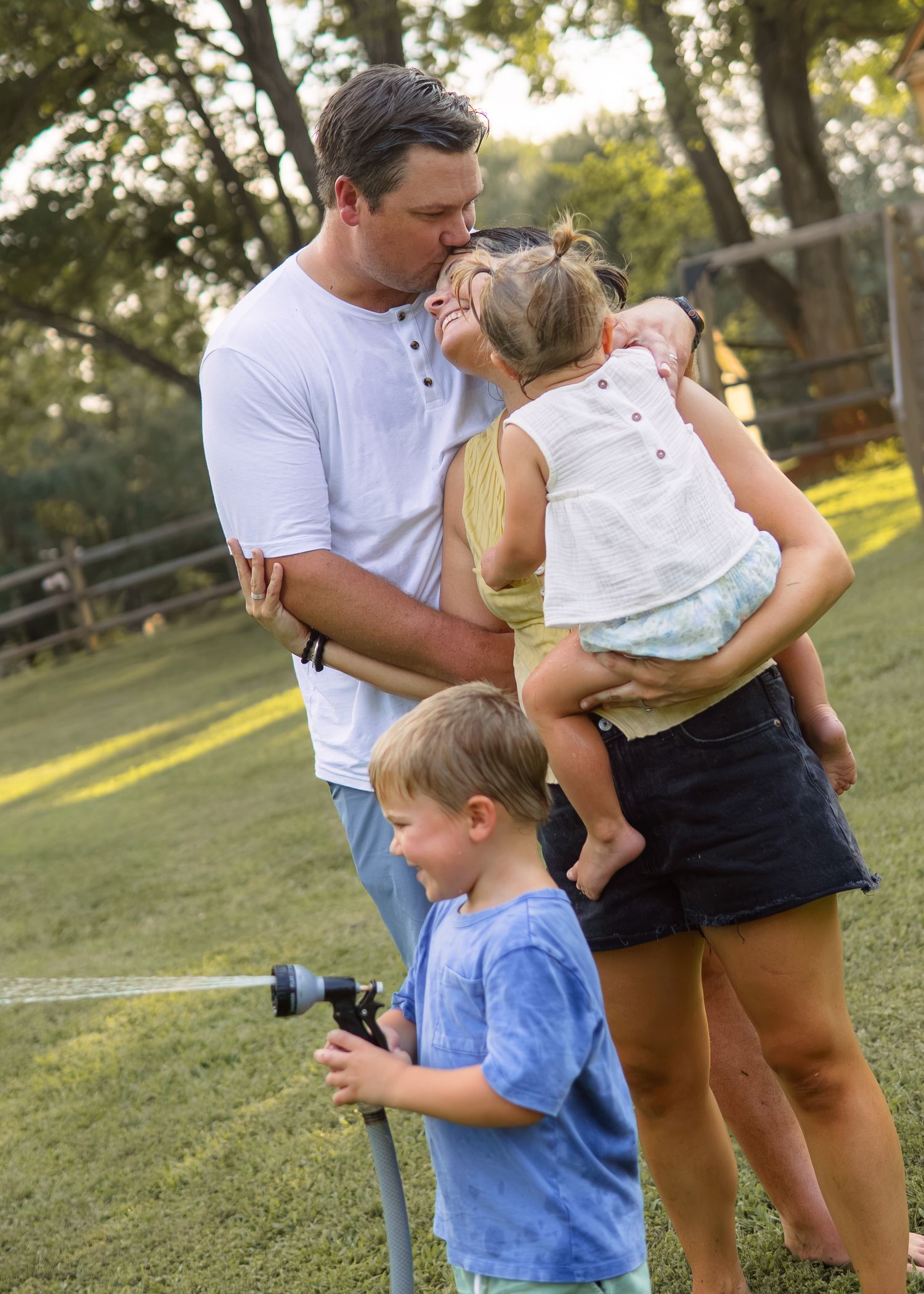 A father kisses his wife while she holds their toddler as their son plays with a garden hose.
