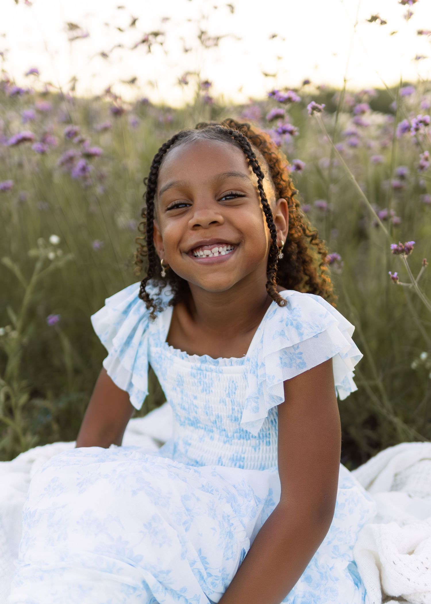 Young Black girl with curly braided hair smiling joyfully in white blue dress among purple wildflowers.