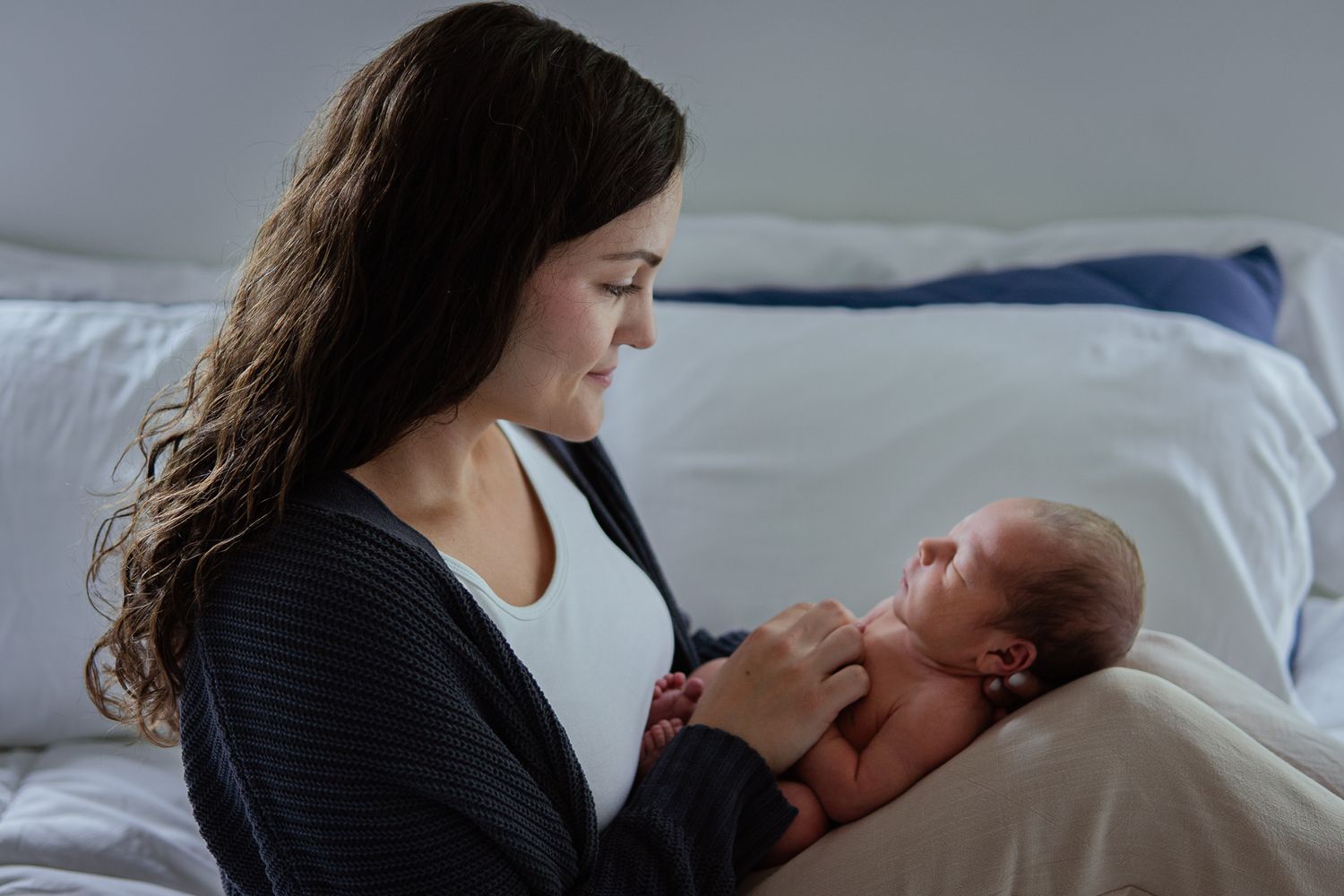 Mother in gray cardigan holding sleeping newborn baby on white bed.