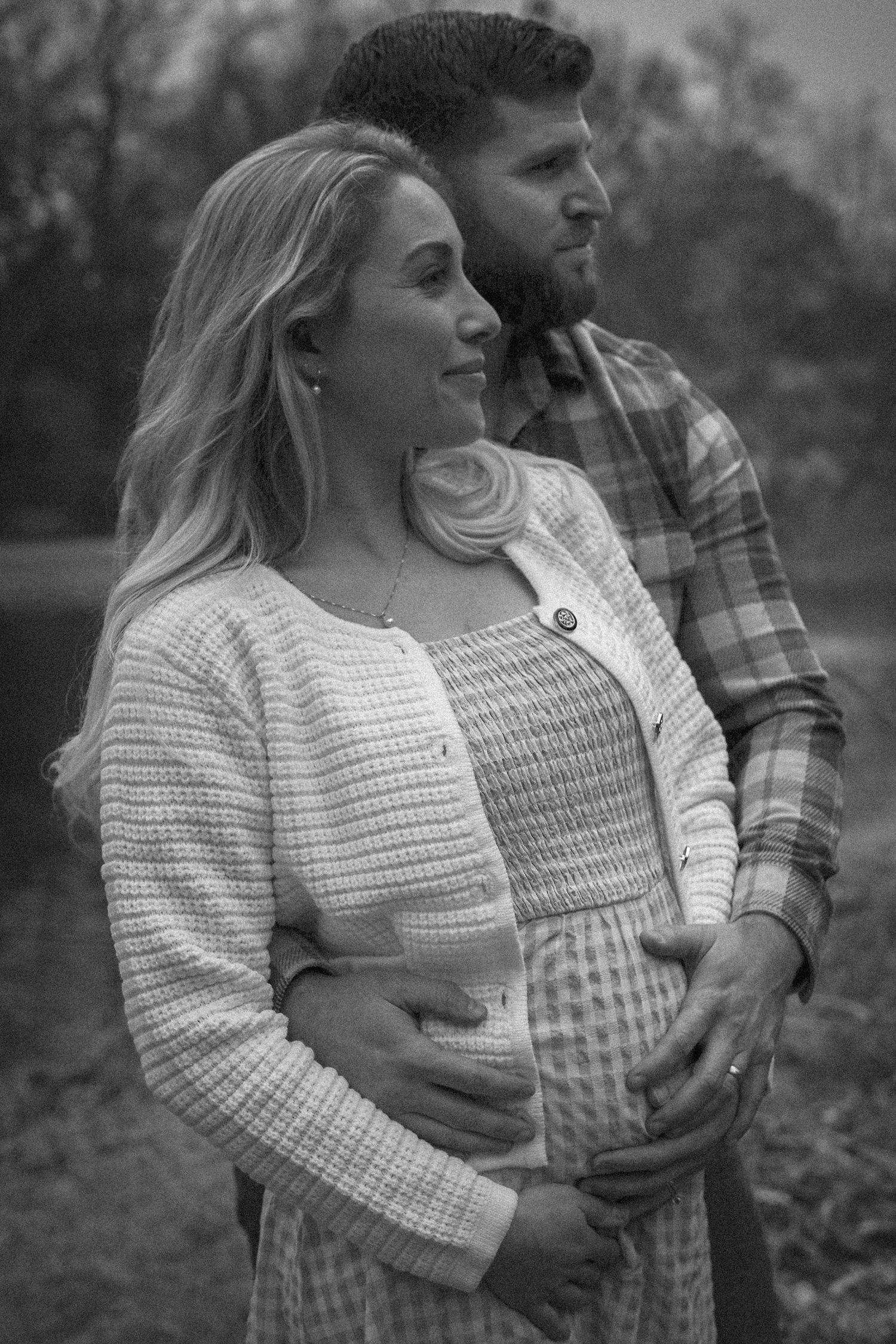 Black and white romantic photo of couple embracing outdoors, with her in knit sweater and him in plaid shirt.