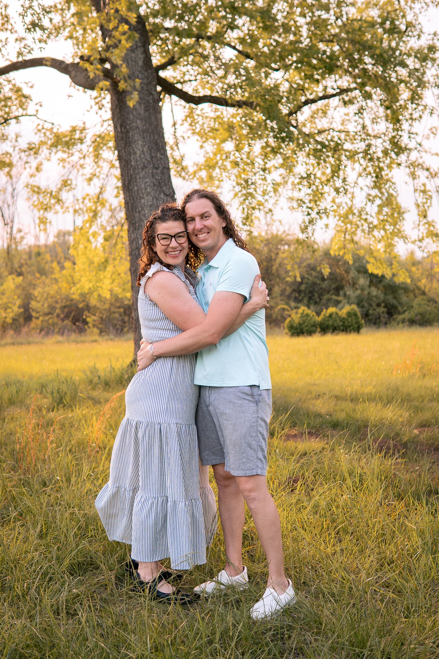 A couple embraces while standing in a sunlit meadow beneath a large tree during golden hour.