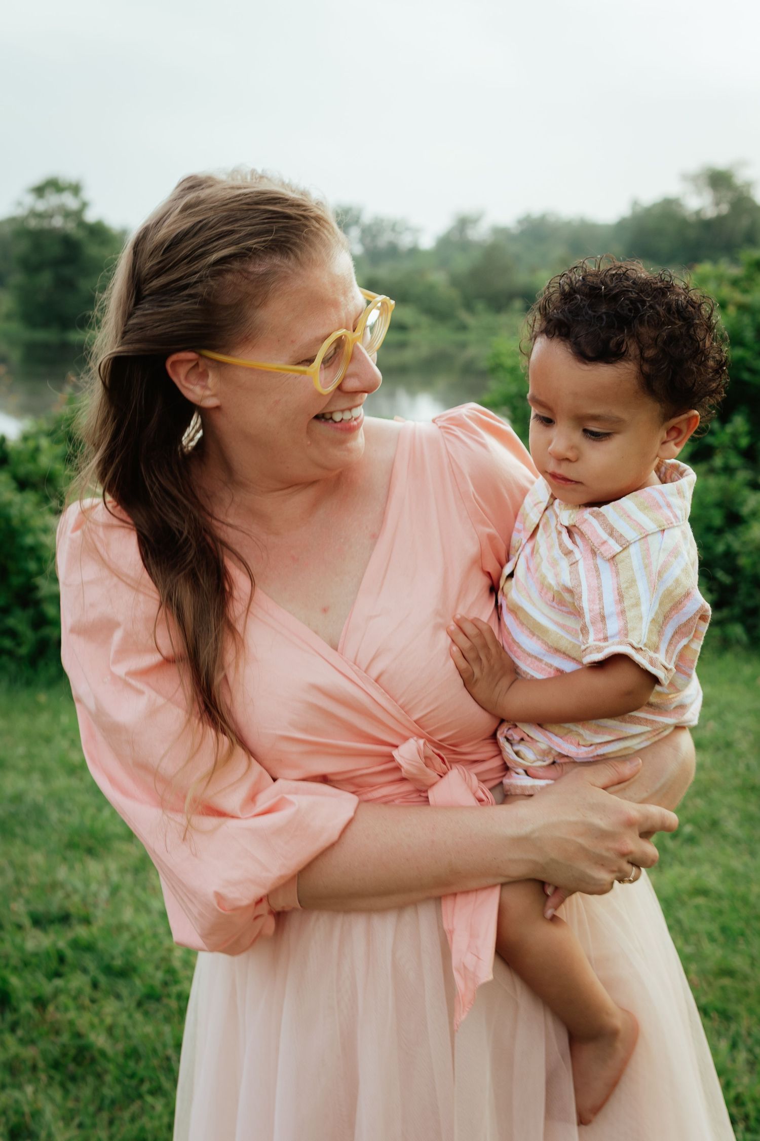 Parent in pink dress holds toddler while smiling outdoors in natural setting.