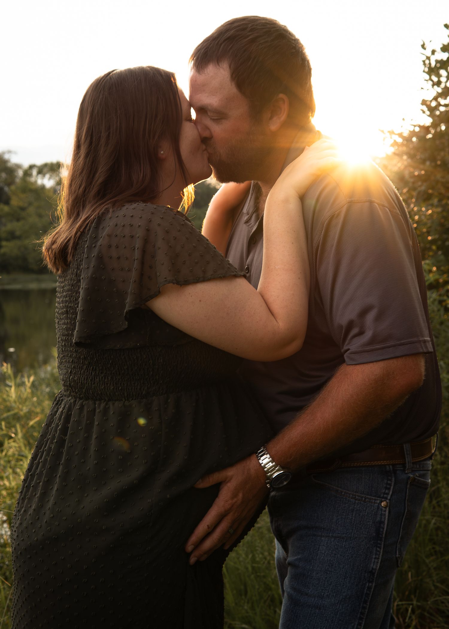 Couple sharing a romantic kiss at golden hour sunset near a pond surrounded by lush greenery.