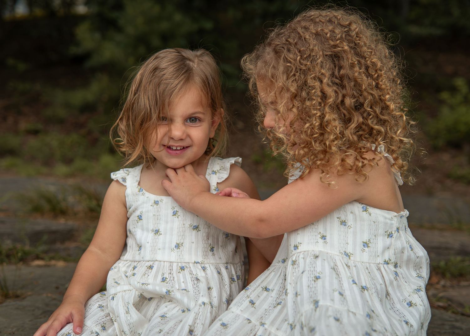 Two young girls in matching white dresses sharing a sweet moment outdoors.