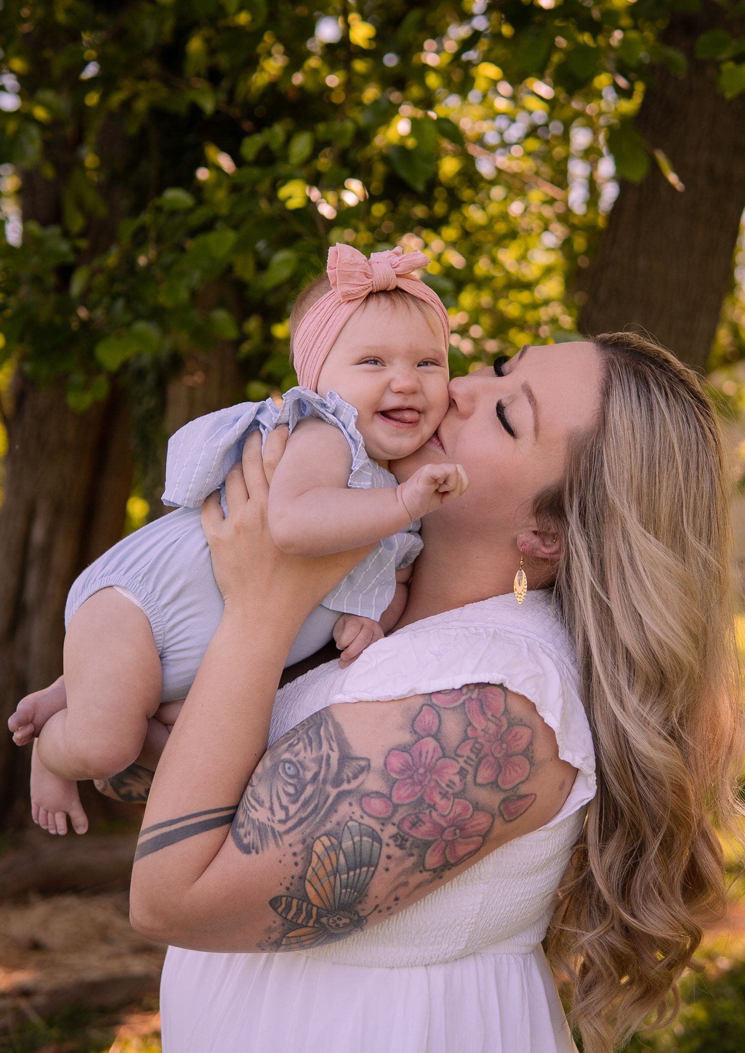 Parent with visible tattoos holds and plays with smiling baby outdoors.