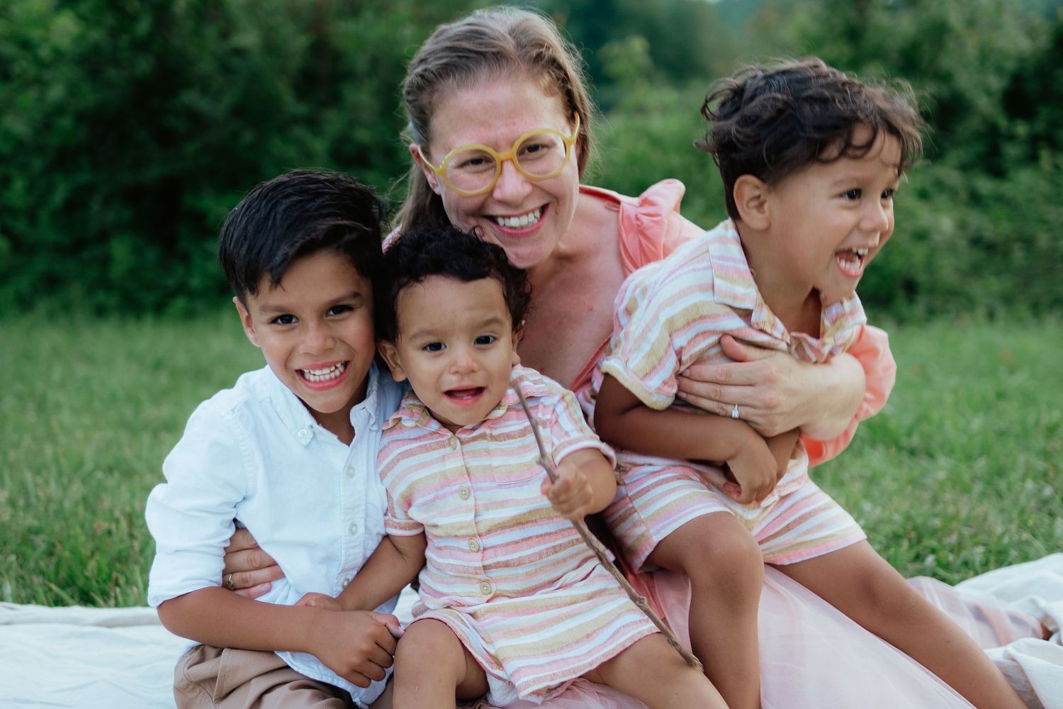 A family with three young children sits together smiling on a blanket outdoors in a grassy area.