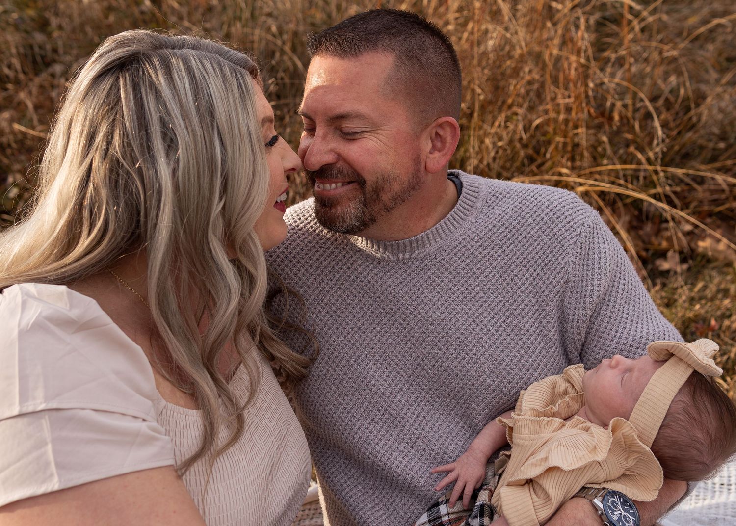 Couple sharing tender moment smiling at newborn baby while sitting in golden field.