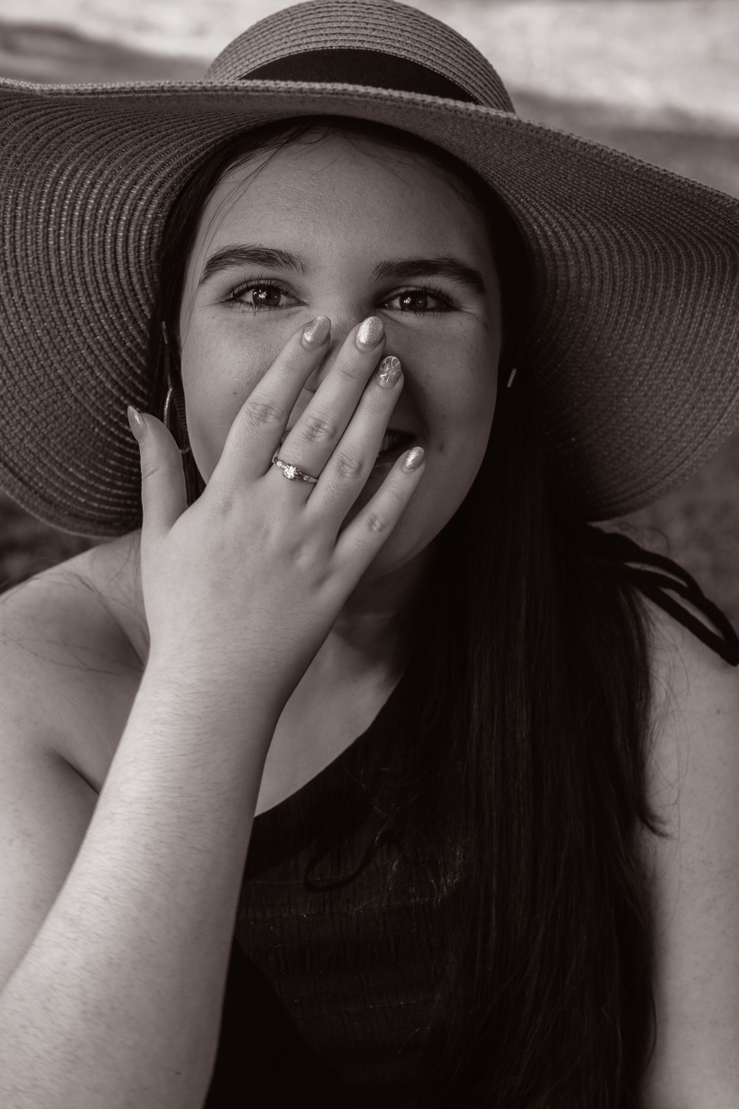 Black and white portrait of smiling young woman in wide brim sun hat covering mouth with ringed hand.