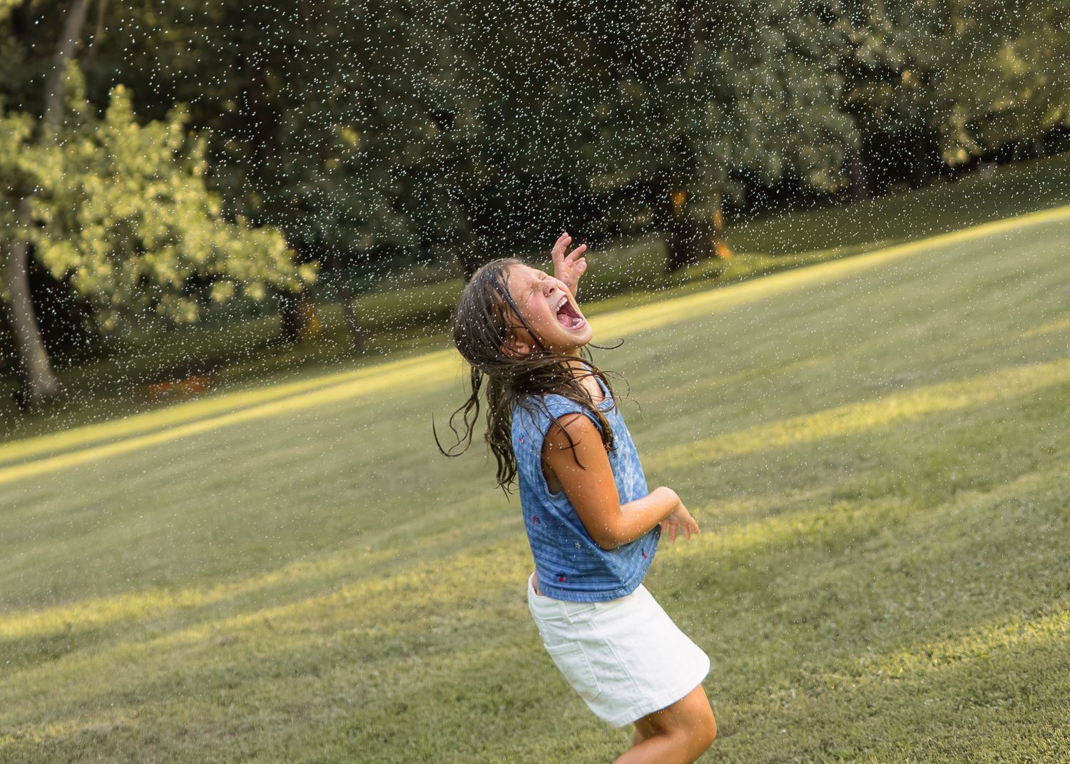 Joyful girl playing in water sprinkler on a sunny green lawn.