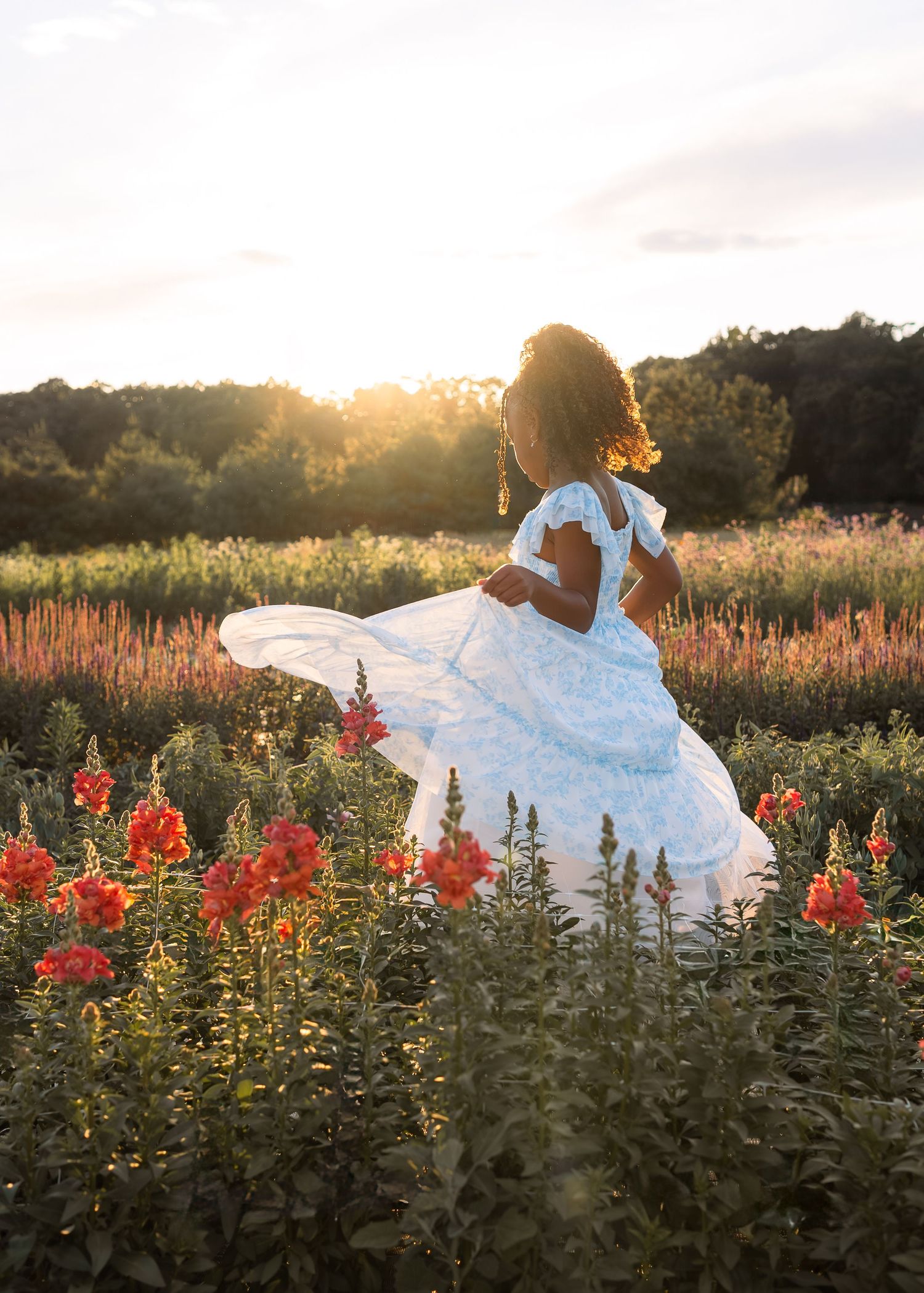 A young Black girl in a white floral dress twirls in a vibrant wildflower field at golden sunset.