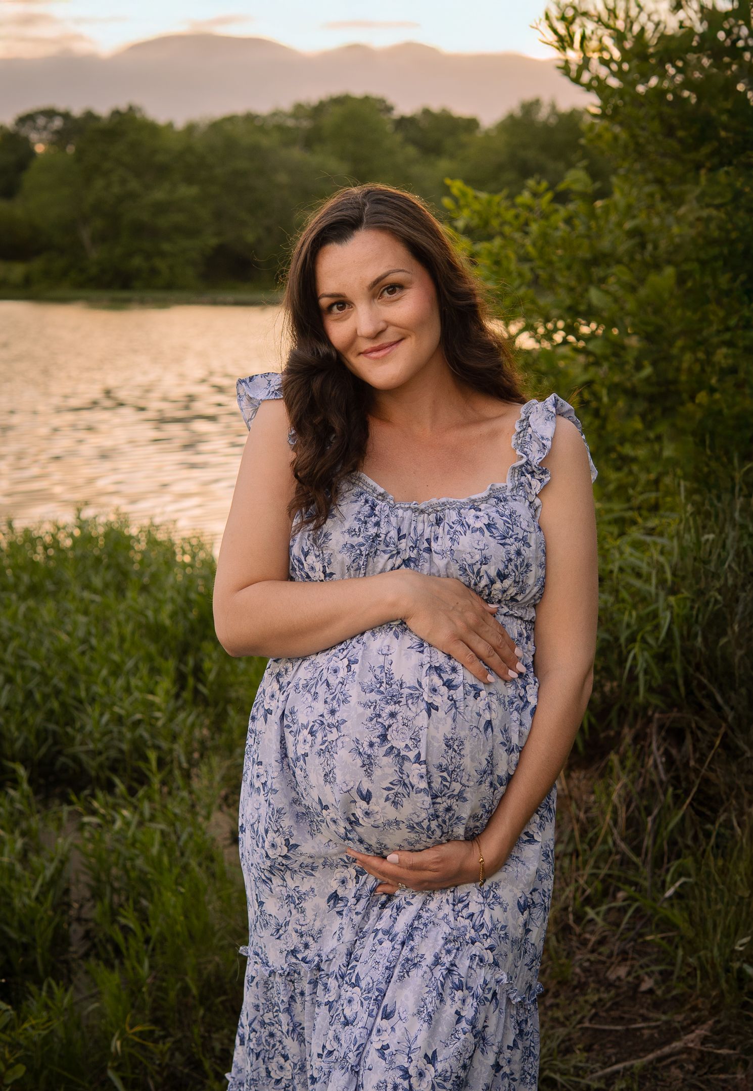 A pregnant woman in a floral sundress stands at sunset near water and greenery in a natural outdoor setting.