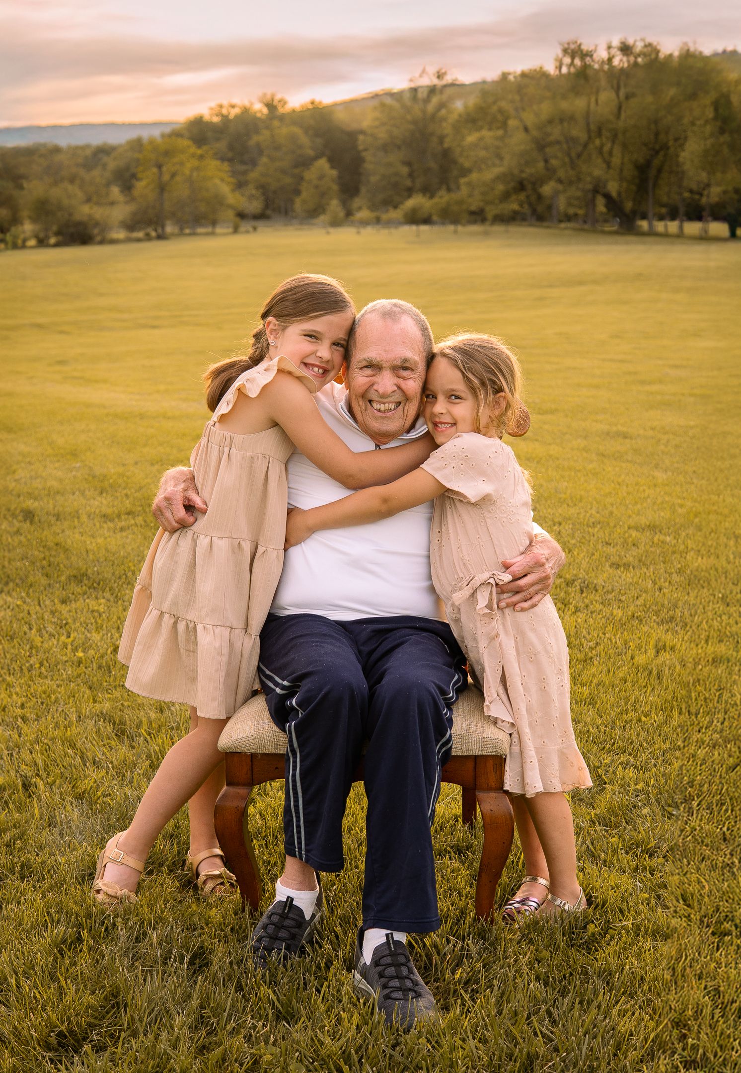 Two children embrace an adult sitting in a chair on a grassy field during a warm sunset.