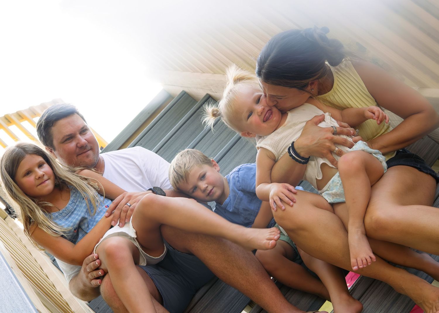 Happy family of five sitting together on wooden steps, laughing and embracing during an outdoor photo session.