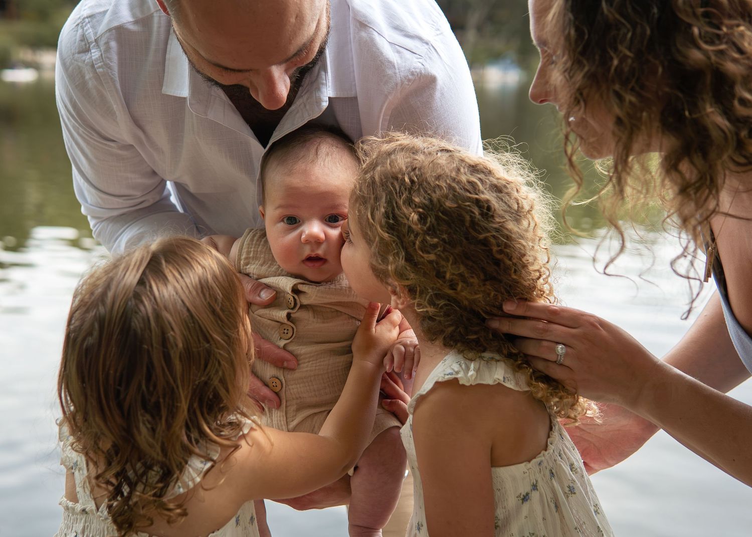 Two sisters kissing a newborn baby held by parents near a serene lake in a heartwarming family portrait.