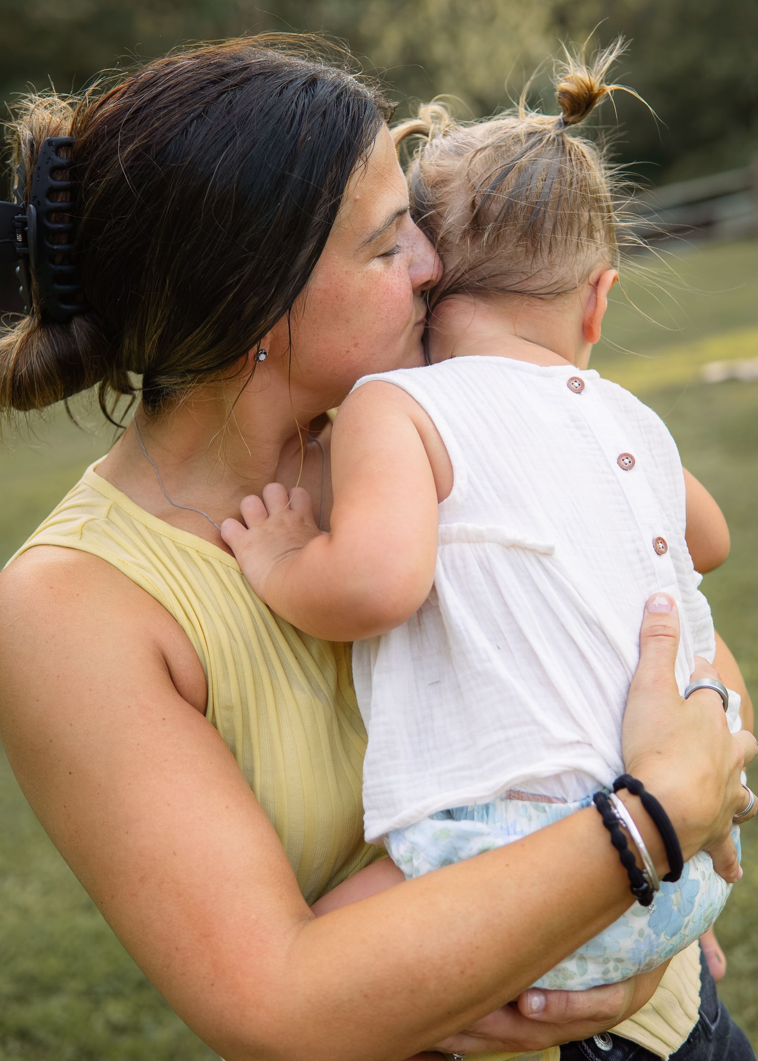 A mother tenderly kisses her toddler daughter on the head while holding her outdoors in a green field.