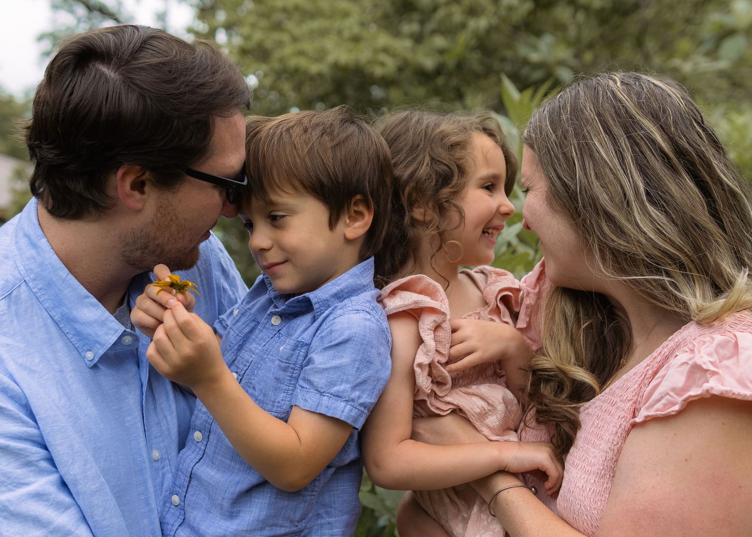 A happy family of four smiling and laughing together outdoors, parents holding young children in a lush green setting.