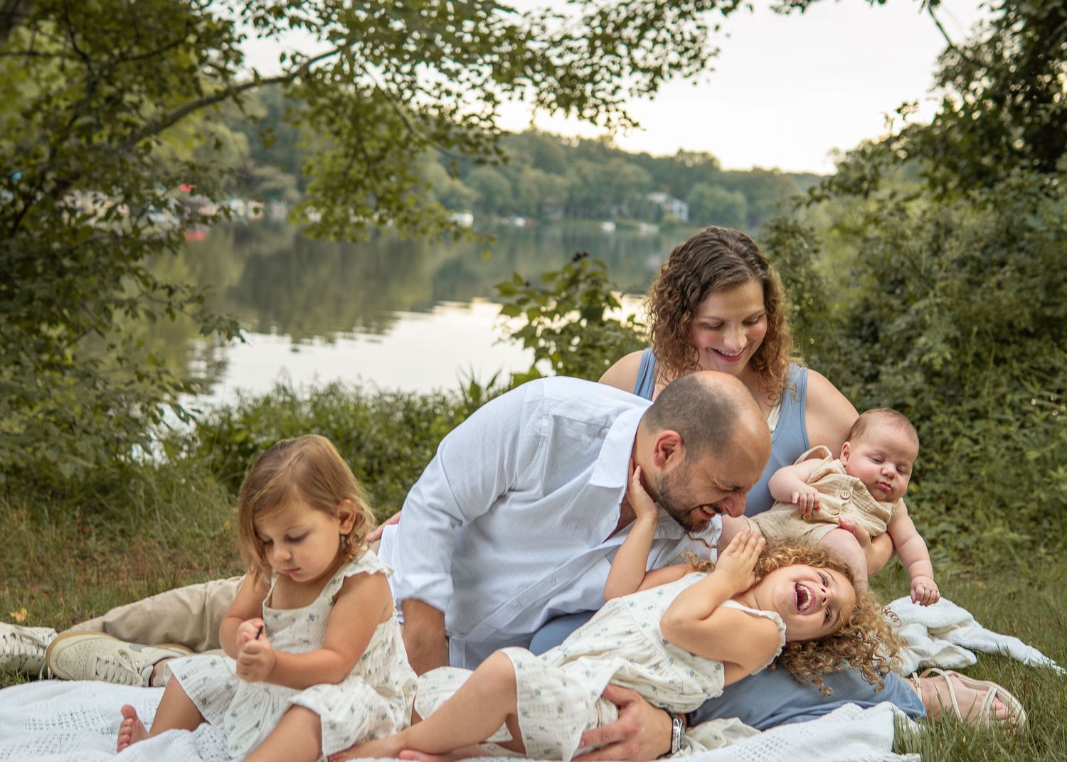 Family of five relaxing on a blanket by a lake surrounded by lush green trees.