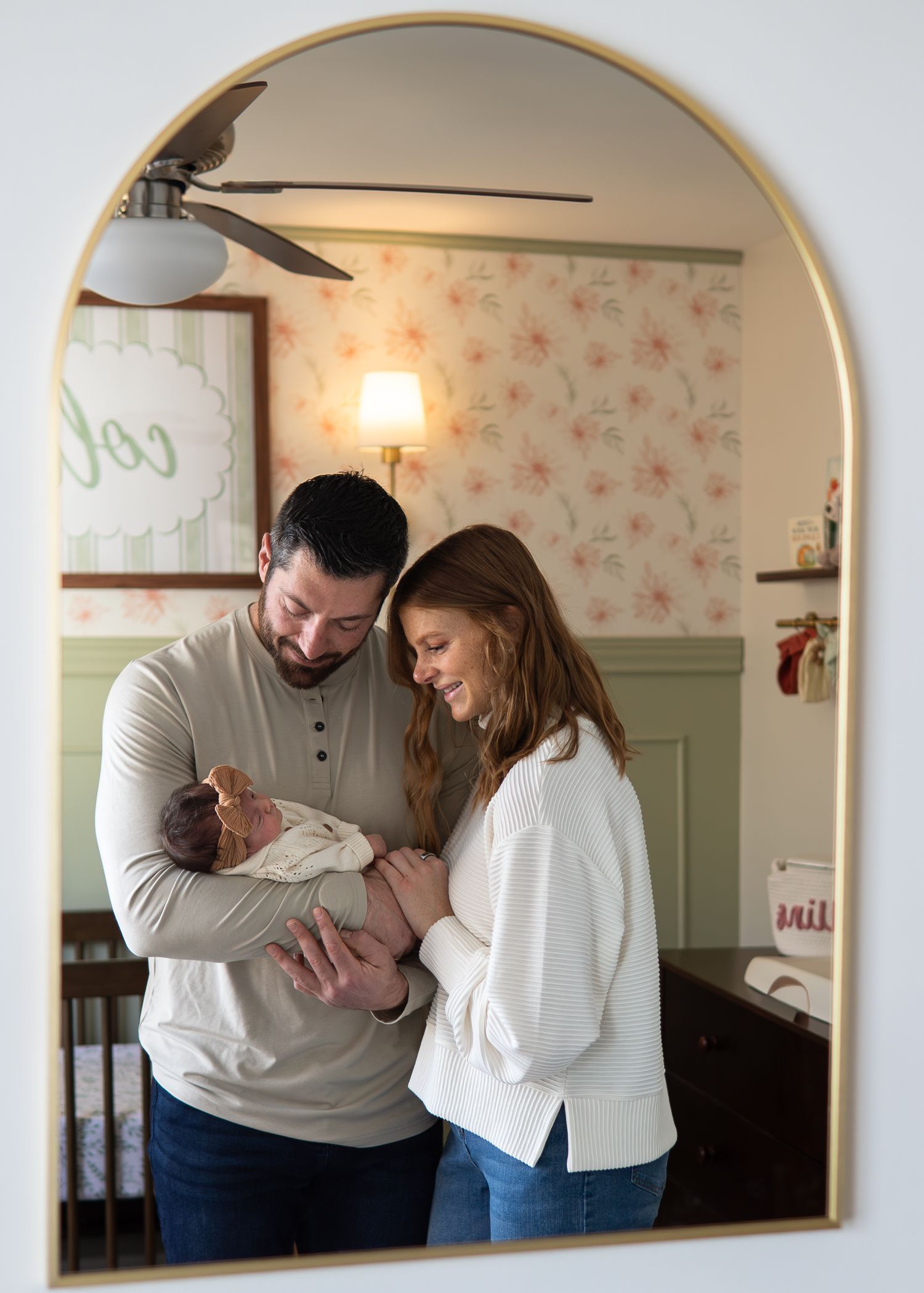 New parents smiling at their newborn baby reflected in an arched gold mirror in nursery.