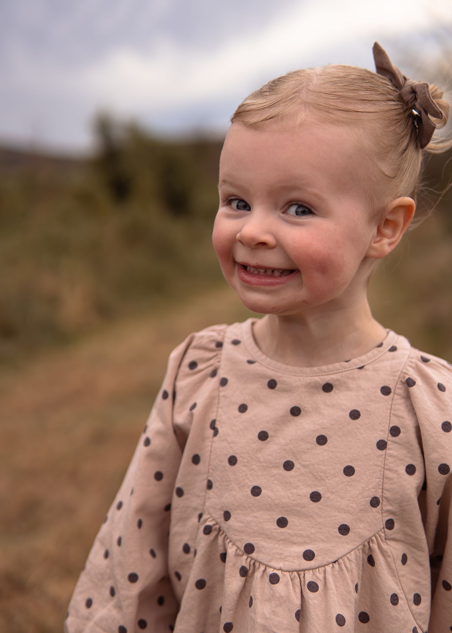 Adorable smiling toddler girl in polka dot dress with hair bow outdoors in nature.