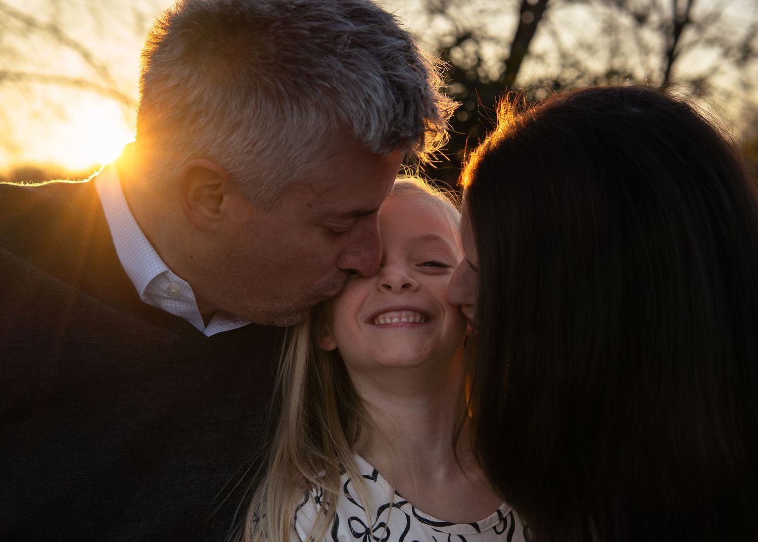 Parents kissing their smiling daughter on the cheeks at golden sunset outdoors.