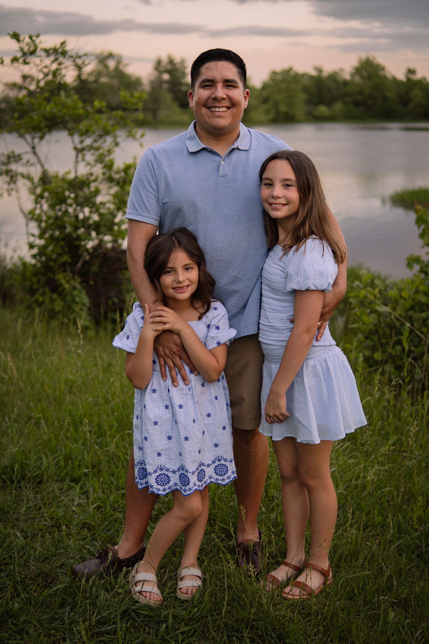 A family of three poses together by a lake during golden hour, wearing coordinating blue and white outfits.