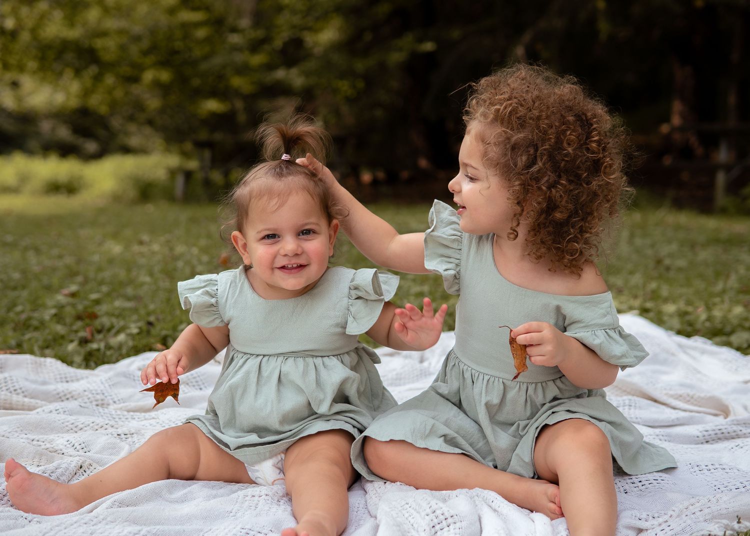 Two young sisters in matching sage green outfits playing together on a blanket.