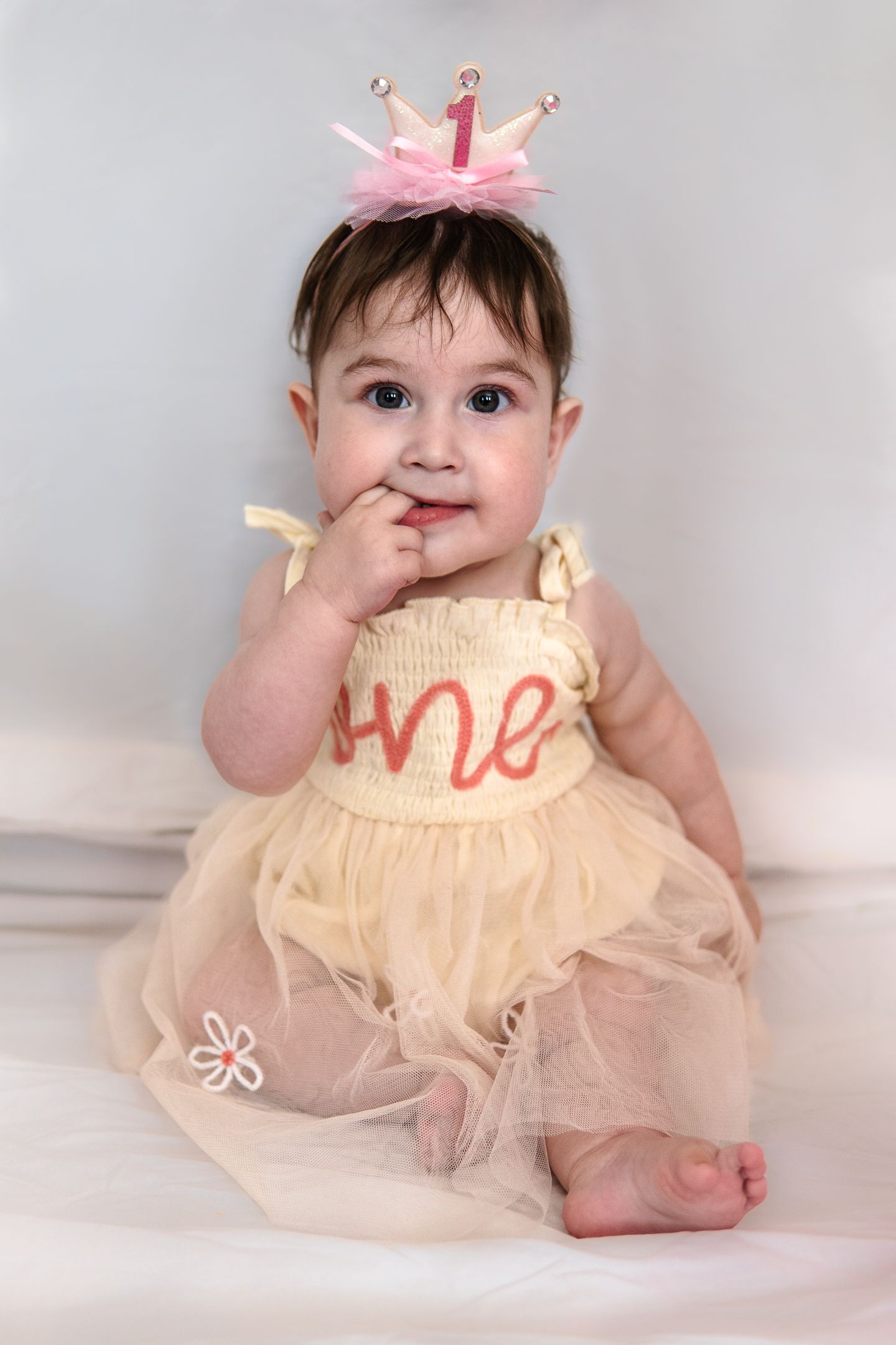 Baby wearing a pink crown and cream tulle dress with finger in mouth during first birthday photoshoot.