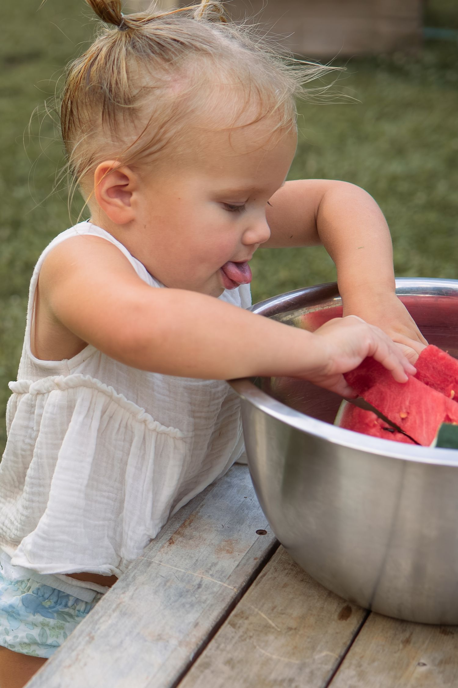 Toddler girl reaching into a metal bowl of watermelon slices on an outdoor wooden table.